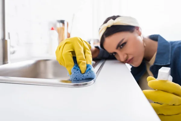 Éponge en main de la femme au foyer sur fond flou lavage évier de cuisine — Photo de stock