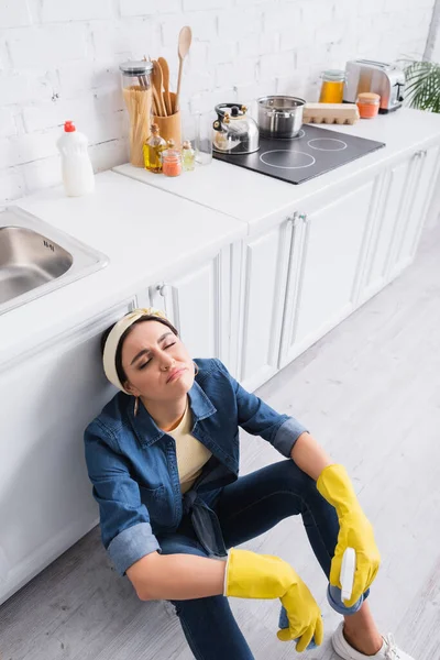 Ama de casa cansada con detergente y esponja sentada en el suelo de la cocina - foto de stock
