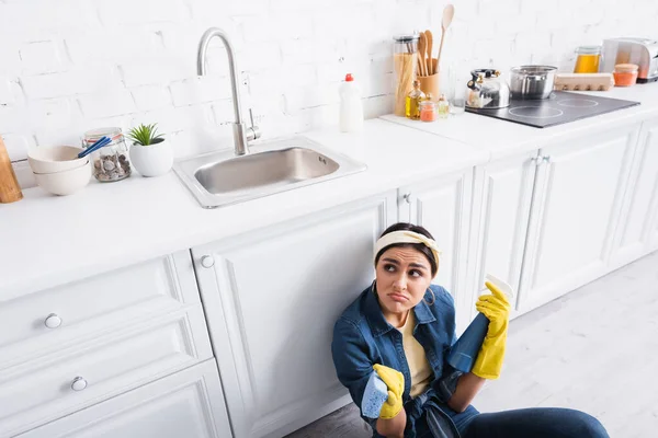 Mulher descontente em luvas de borracha segurando garrafa detergente e esponja na cozinha — Fotografia de Stock