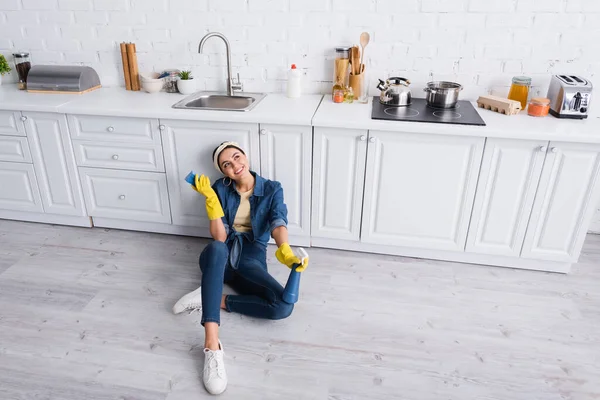 Happy housewife with detergent and sponge sitting on floor in kitchen — Stock Photo