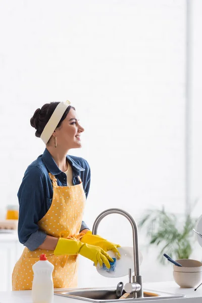Веселая женщина в фартуке и резиновых перчатках моет тарелку на кухне — стоковое фото