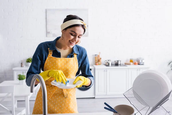 Mulher positiva em placa de lavagem avental na cozinha — Fotografia de Stock