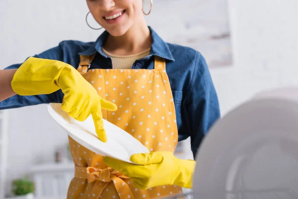 Vista ritagliata di donna sorridente in guanti di gomma che tiene piatto pulito — Foto stock