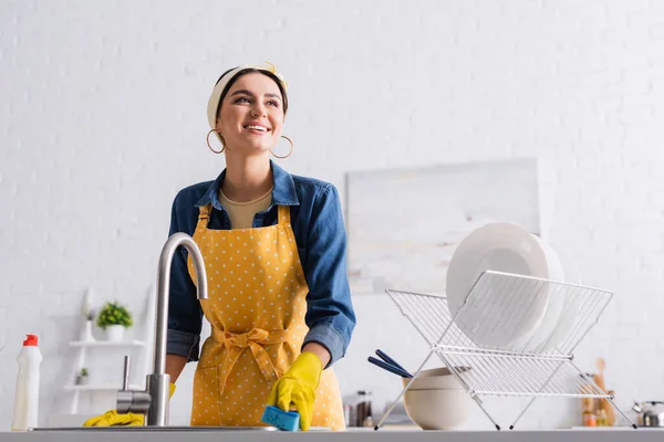 Sorrindo dona de casa em luvas de borracha segurando esponja perto utensílio e pia — Fotografia de Stock