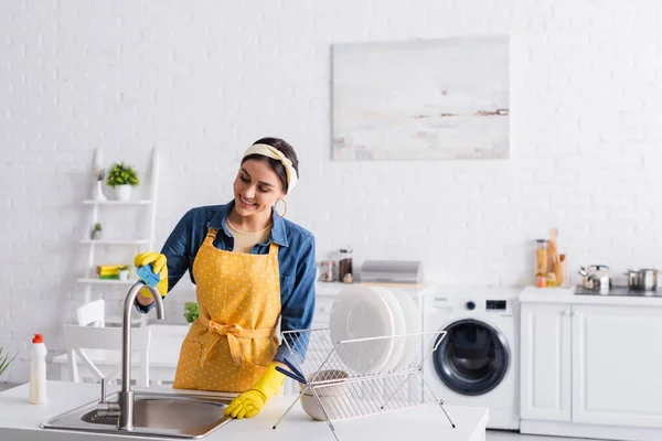 Улыбающаяся домохозяйка чистит кран возле тарелок на кухне — стоковое фото