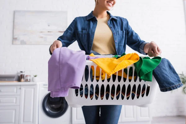 Vista recortada de la cesta con ropa en manos de ama de casa borrosa en la cocina - foto de stock