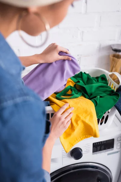 Mulher desfocada segurando roupas perto de cesta e máquina de lavar roupa — Fotografia de Stock