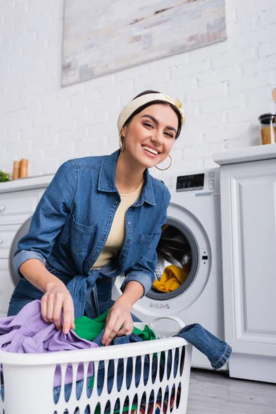 Ama de casa sonriendo cerca de la cesta con ropa y lavadora en casa - foto de stock