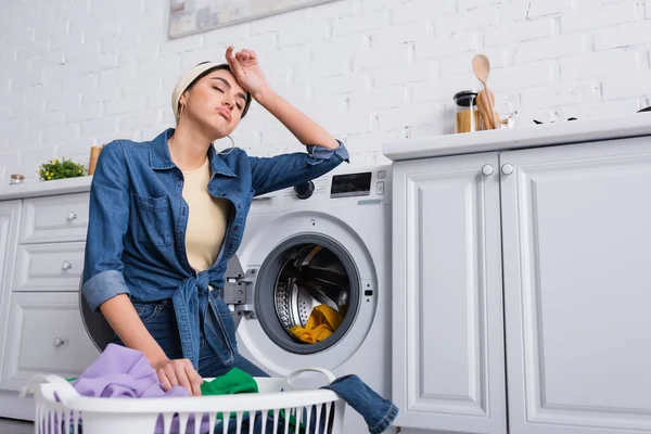 Tired housewife near basket with clothes and washing machine in kitchen — Stock Photo