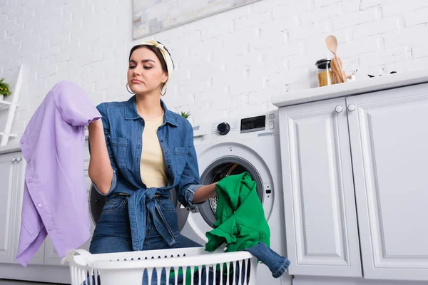 Dona de casa olhando para roupas perto de cesta e máquina de lavar roupa — Fotografia de Stock