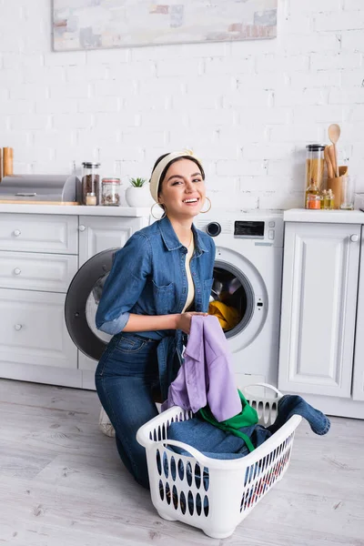 Casalinga positiva che guarda la macchina fotografica vicino al cesto con lavanderia e lavatrice in cucina — Foto stock