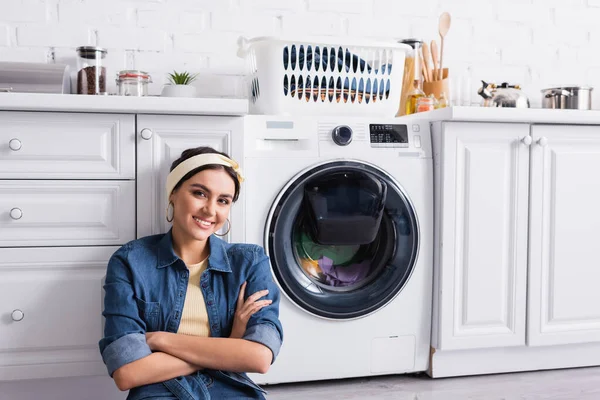 Улыбающаяся домохозяйка смотрит в камеру возле стиральной машины на кухне — стоковое фото