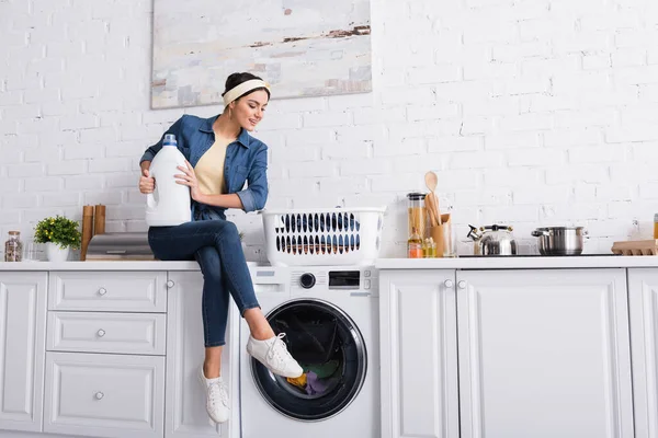 Dona de casa alegre com detergente sentado perto da máquina de lavar roupa e roupas — Fotografia de Stock