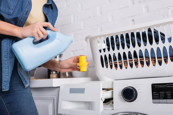 Cropped view of housewife holding cap and washing liquid near basket and washing machine — Stock Photo
