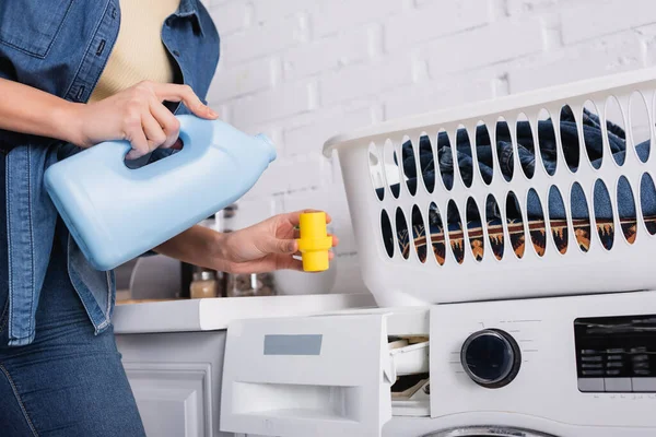 Vue recadrée de la femme au foyer tenant le chapeau et le liquide de lavage près du panier et de la machine à laver — Photo de stock