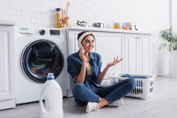 Femme au foyer souriante parlant sur smartphone près de détergent et lave-linge dans la cuisine — Photo de stock