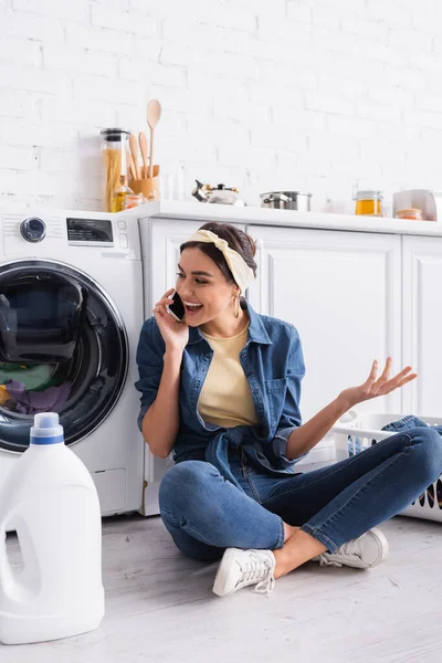 Femme au foyer positive parlant sur smartphone près de détergent et machine à laver — Photo de stock