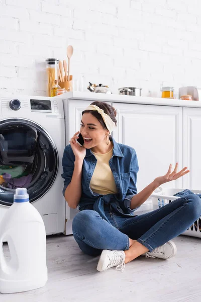 Ama de casa positiva hablando en el teléfono inteligente cerca de detergente y lavadora - foto de stock