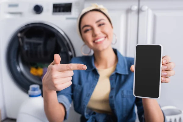 Dona de casa desfocada apontando para smartphone perto da máquina de lavar roupa — Fotografia de Stock
