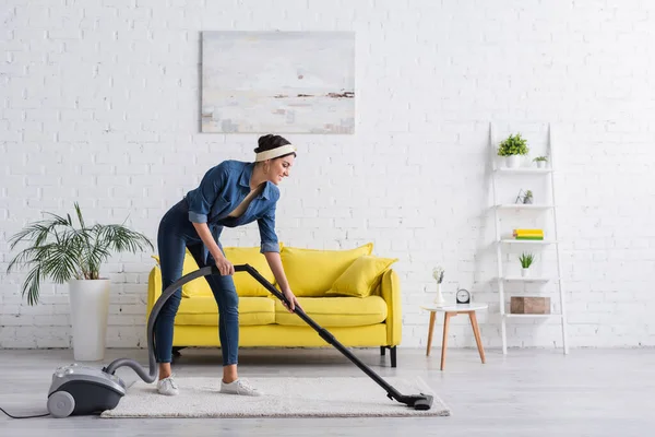 Junge Frau lächelt und putzt Teppich mit Staubsauger — Stockfoto