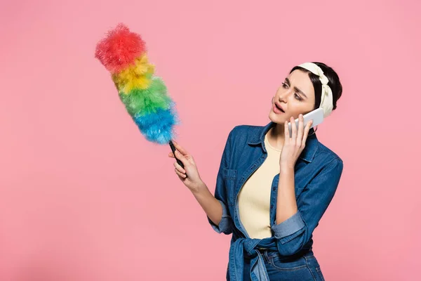 Housewife talking on smartphone and holding dust brush isolated on pink — Stock Photo