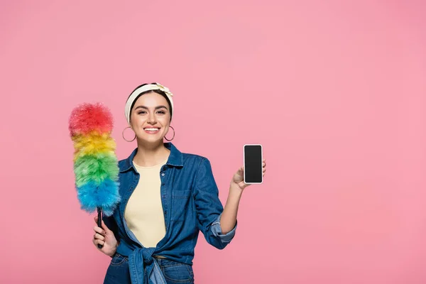 Femme au foyer souriante avec brosse à poussière tenant téléphone portable avec écran blanc isolé sur rose — Photo de stock
