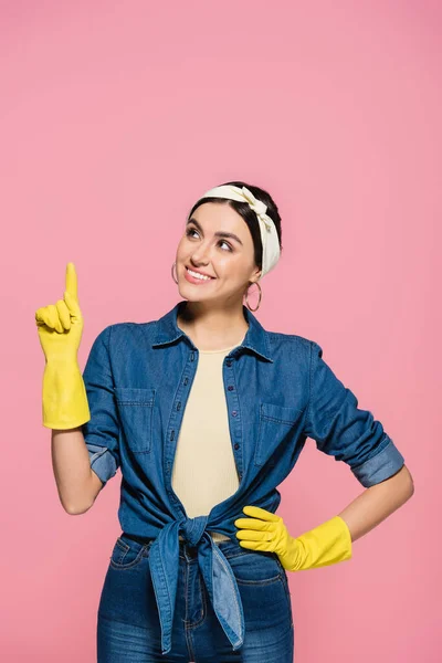 Femme au foyer souriante en gants de caoutchouc pointant vers le haut avec doigt isolé sur rose — Photo de stock