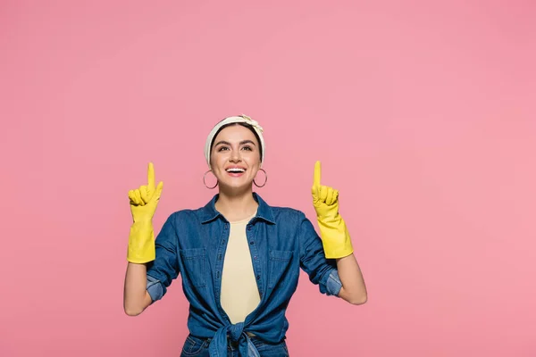 Femme au foyer positive dans des gants en caoutchouc pointant vers le haut avec les doigts isolés sur rose — Photo de stock