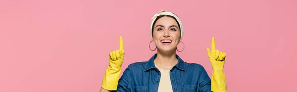 Улыбающаяся домохозяйка в жёлтых резиновых перчатках, указывающая пальцами, изолированными на розовом, баннер — стоковое фото