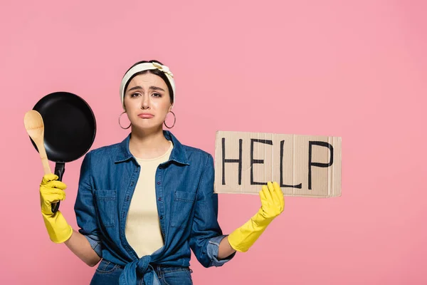 Sad housewife in rubber gloves holding frying pan and placard with help lettering isolated on pink — Stock Photo