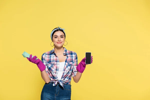 Веселая домохозяйка держит тряпку и смартфон изолированными на желтый — стоковое фото