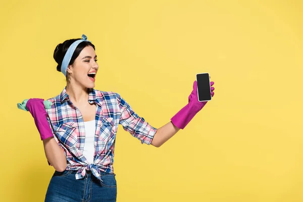 Ama de casa positiva sosteniendo trapo y teléfono inteligente con pantalla en blanco aislado en amarillo - foto de stock
