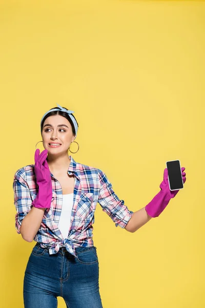 Confused housewife in rubber gloves holding smartphone with blank screen isolated on yellow — Stock Photo