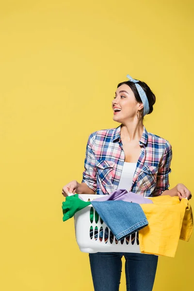 Cheerful housewife holding basket with clothes isolated on yellow — Stock Photo