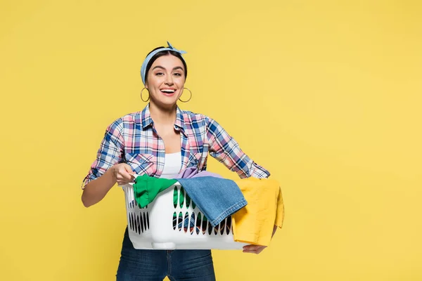 Young woman holding basket with clothes and smiling at camera isolated on yellow — Stock Photo