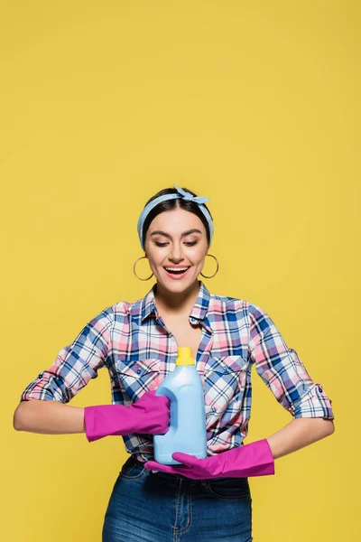 Mulher sorridente em luvas de borracha olhando para garrafa de detergente isolado no amarelo — Fotografia de Stock