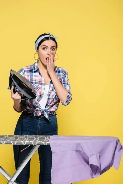 Excited housewife holding iron near board and shirt on yellow background — Stock Photo