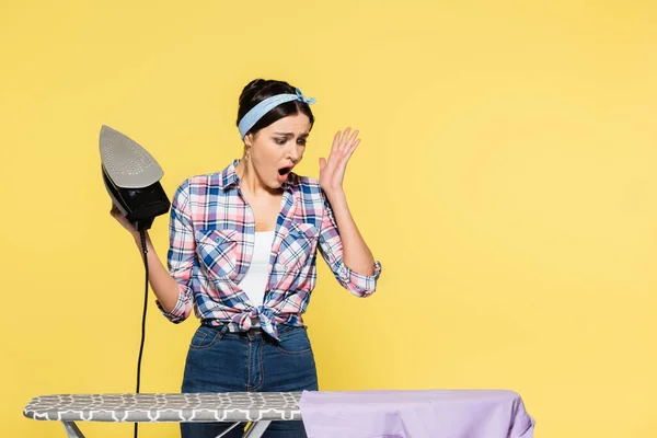 Worried woman holding iron near clothes on board isolated on yellow — Stock Photo