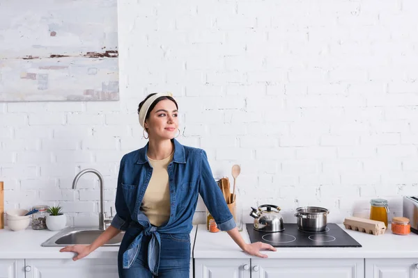 Улыбающаяся домохозяйка в джинсовой рубашке, шлифующая возле кухонного стола — стоковое фото