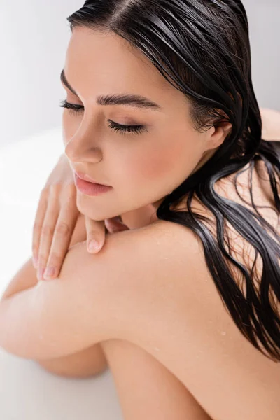 Sensual mujer joven con el pelo mojado y los ojos cerrados sentado en la bañera con leche - foto de stock