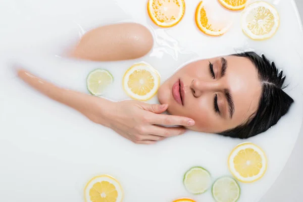 Vue de dessus de la jeune femme touchant le visage tout en se baignant dans le lait avec des tranches d'agrumes — Photo de stock