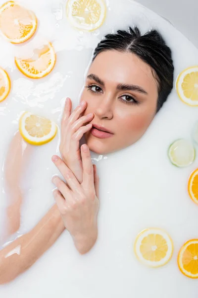 Woman in milk bath with sliced citruses touching face while looking at camera — Stock Photo