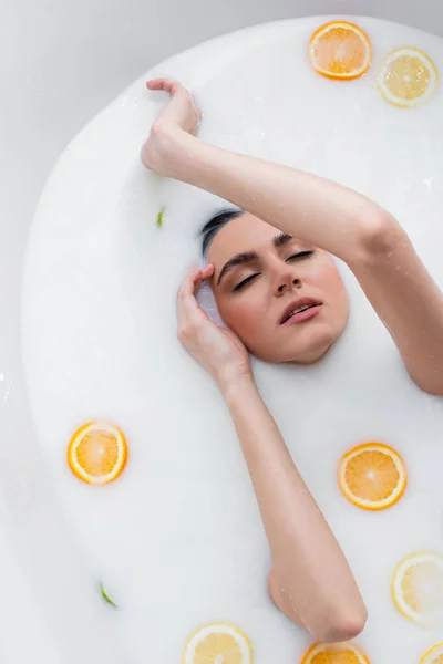 Top view of woman with closed eyes bathing in milk with sliced citruses — Stock Photo