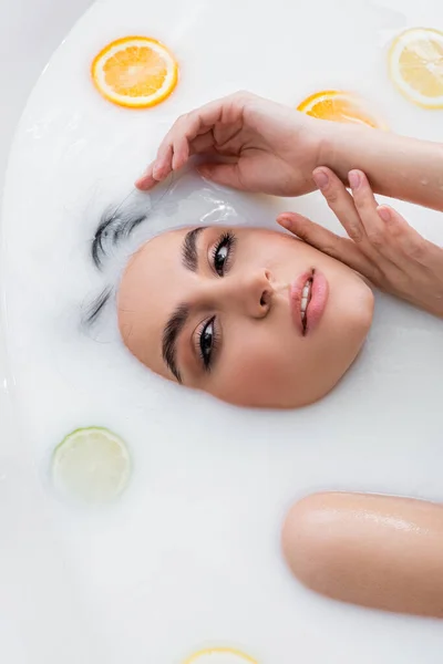 Bonita mujer cogida de la mano cerca de la cara y mirando a la cámara mientras se relaja en el baño de leche con cítricos - foto de stock