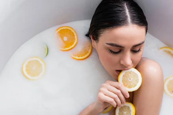 Mulher bonita mordendo fatia de limão fresco enquanto relaxa no banho de leite — Fotografia de Stock