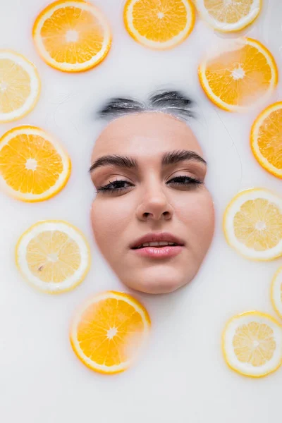 Vista superior de la cara femenina en baño lechoso con limones en rodajas y naranjas - foto de stock