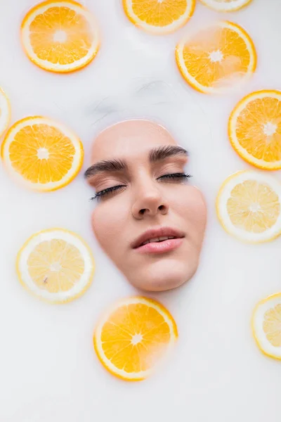 Vue de dessus du visage féminin dans un bain de lait avec du citron et de l'orange tranchés — Photo de stock