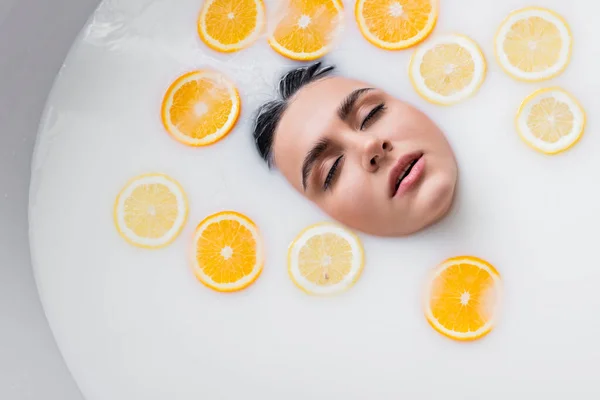 Vista superior de la cara femenina en baño lechoso con limón en rodajas y naranja - foto de stock