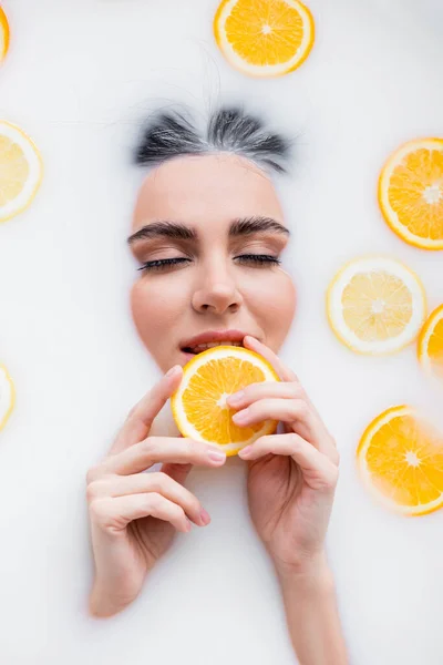 Young woman holding slice of fresh orange while relaxing in milk bath — Stock Photo