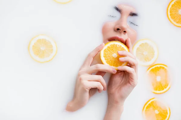 Vista de ángulo alto de la mujer sosteniendo rebanada de naranja mientras sumerge la cara en el baño de leche - foto de stock